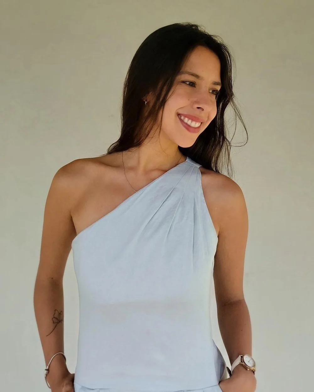 Ludmila Passarini sorrindo, de pé, olhando para o lado, com blusa clara de um ombro só